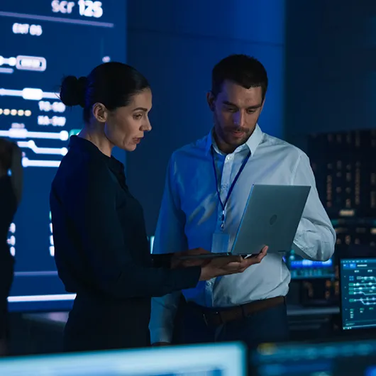 Two cybersecurity professionals analyze data on a laptop in a high-tech operations center, surrounded by multiple monitors and digital displays.