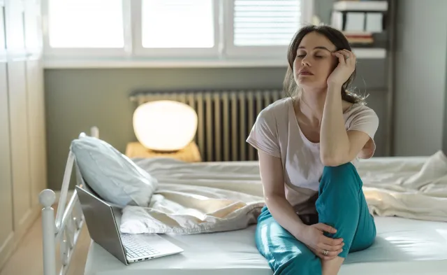 A young woman sitting on her bed in a sunny bedroom, appearing deep in thought and showing signs of tiredness. The scene suggests contemplation, relaxation, or a morning routine in a cozy setting.
